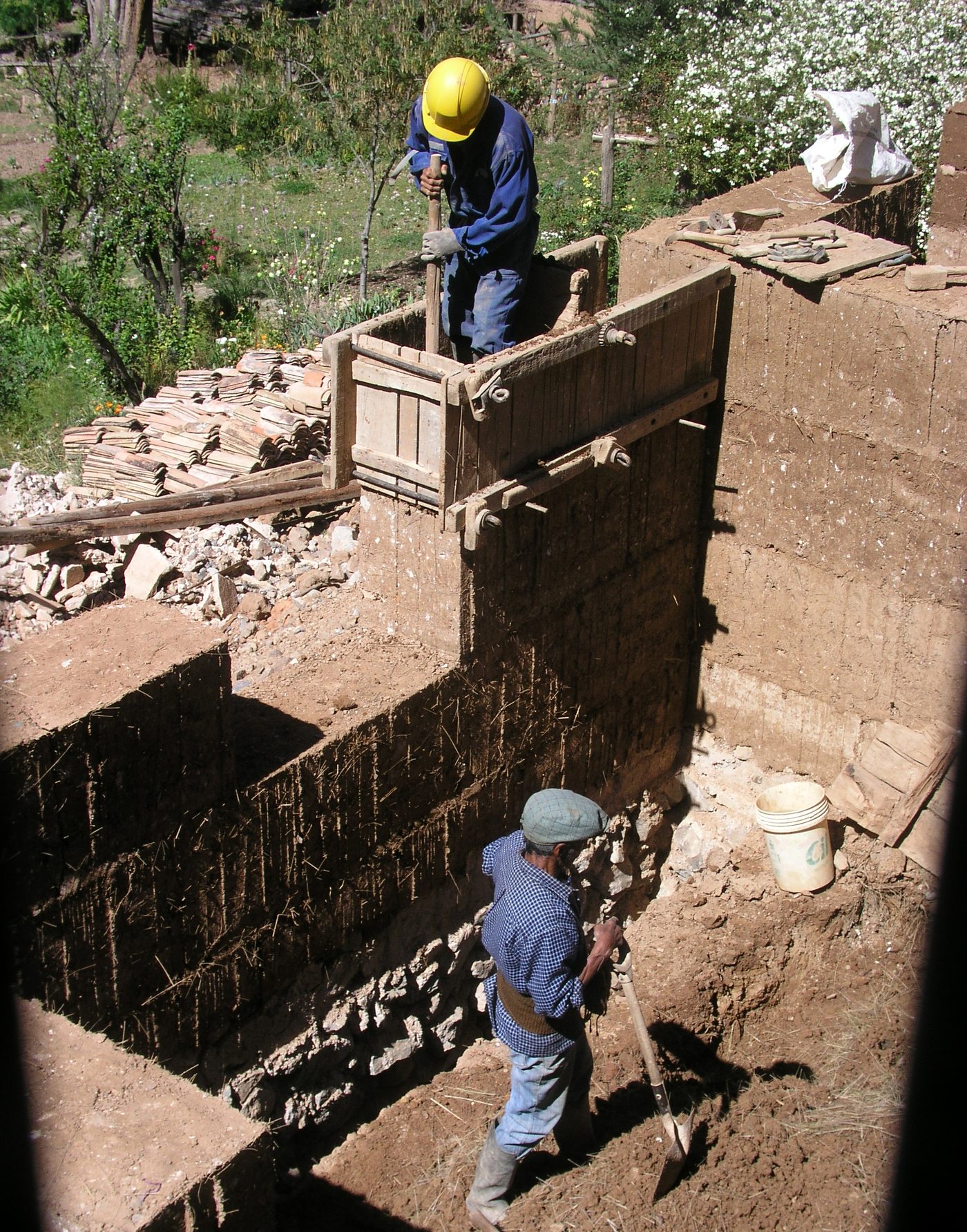 Two builders tamping moist earth inside timber formwork to raise a rammed earth wall